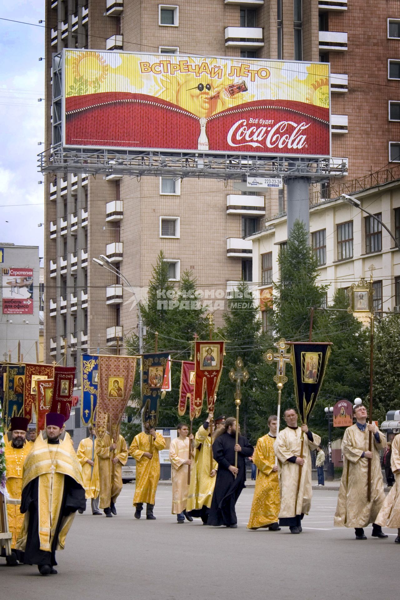 Священнослужители идут по улице города на фоне рекламного плаката с надписью: `Встречай лето`