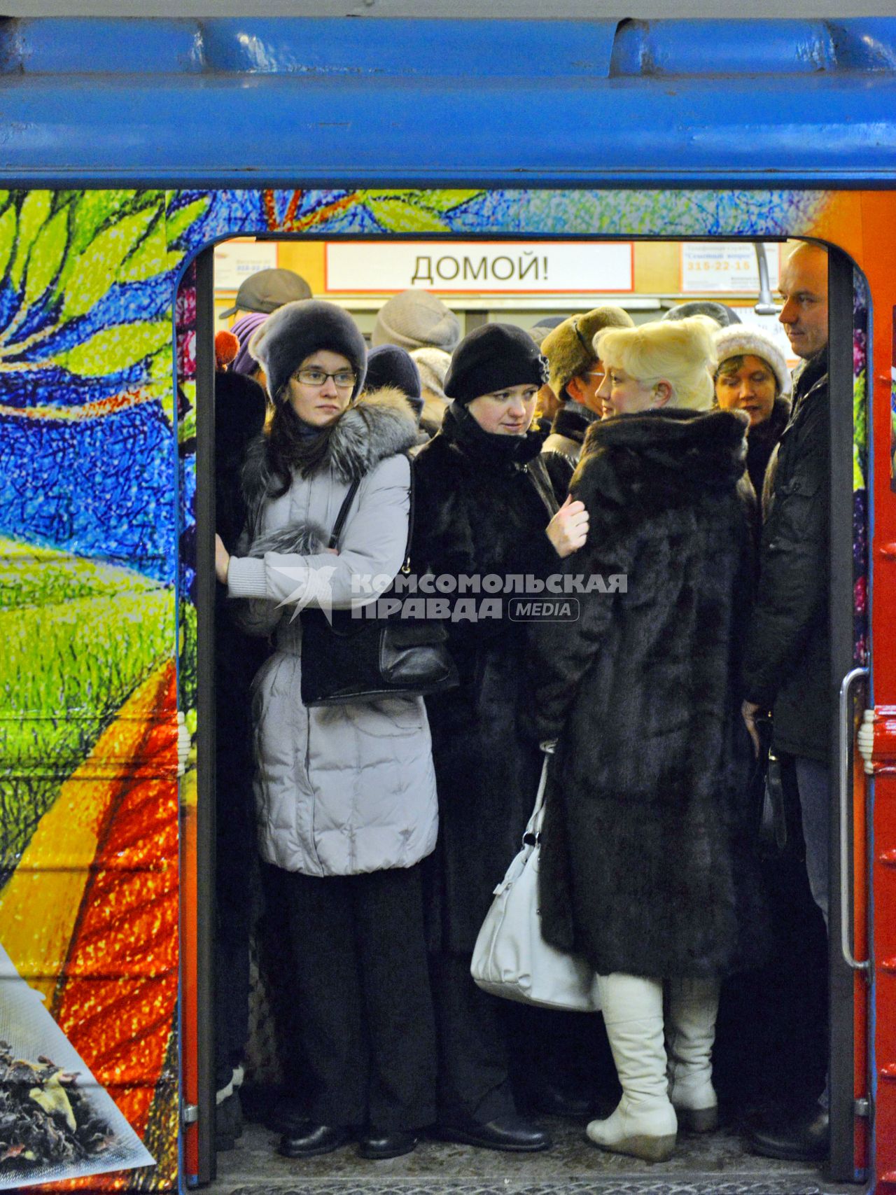 В дверях поезда метро плотно стоят люди в теплой зимней одежде, на заднем плане плакат: `Домой!`