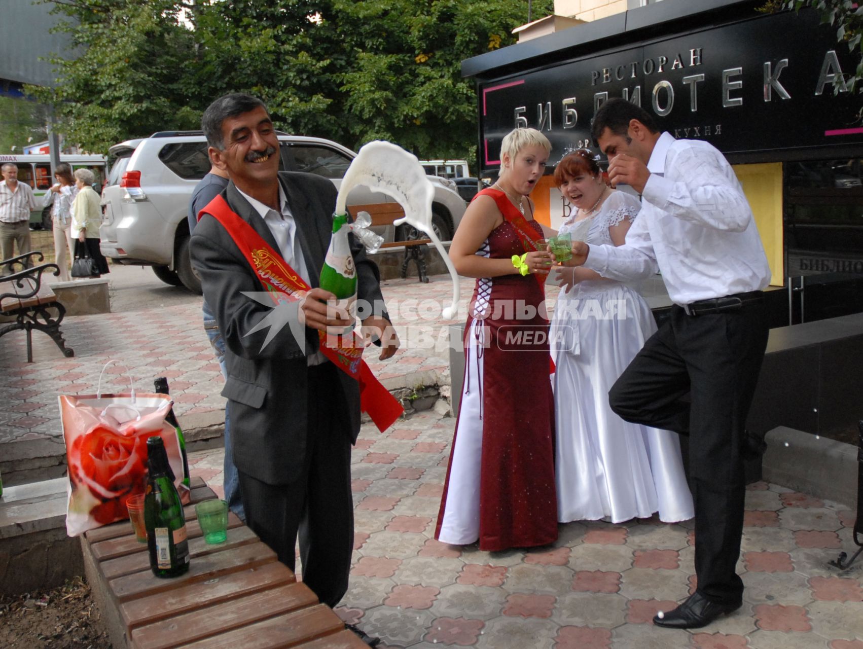Русско-кавказская свадьба. Свидетель кавказец открывает шампанское. Жених кавказец и русские невеста со свидетельницей стоят рядом с бокалами.