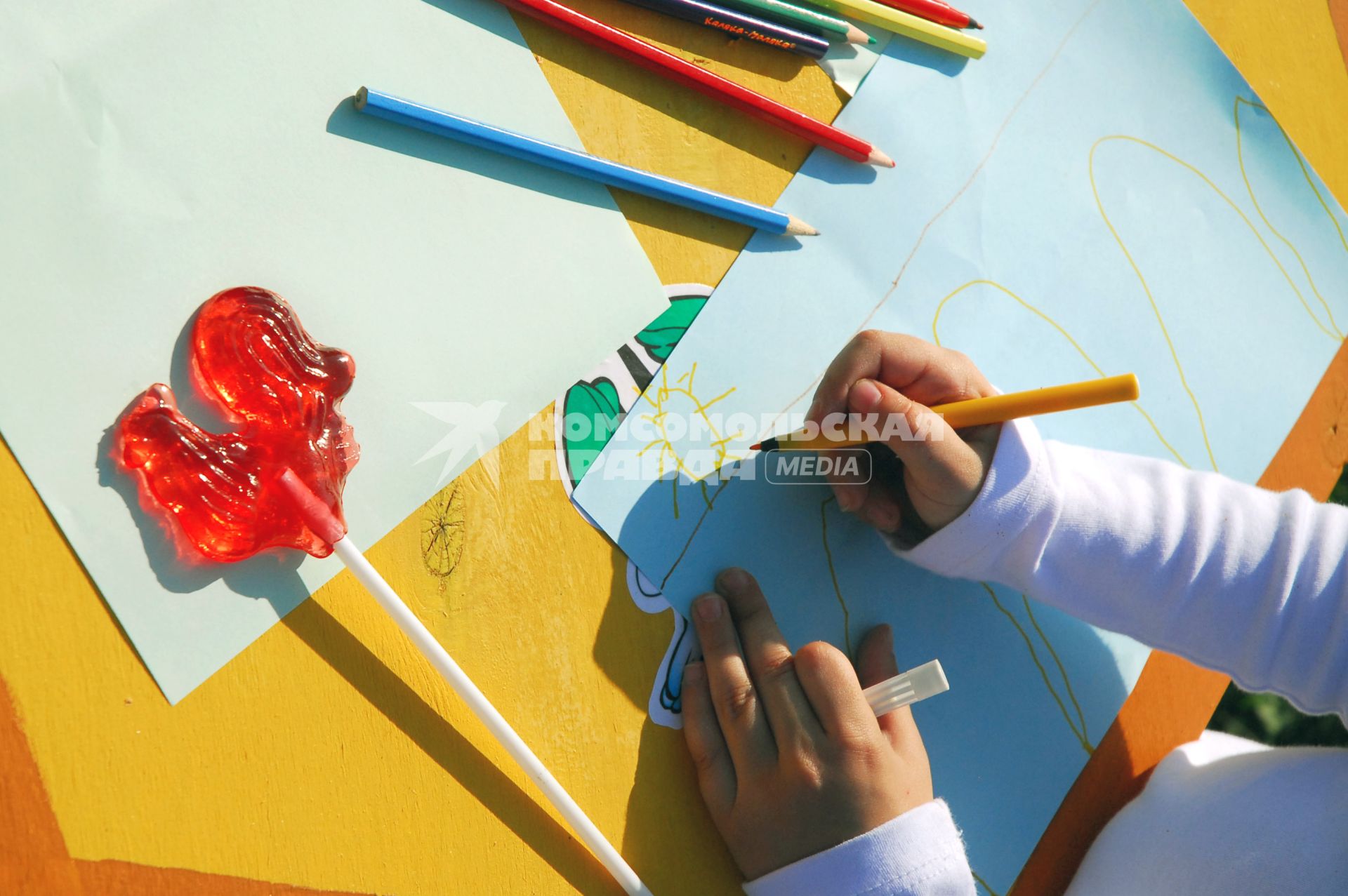 Руки рисующей девочки, цветные карандаши и леденцовый петушок на палочке.