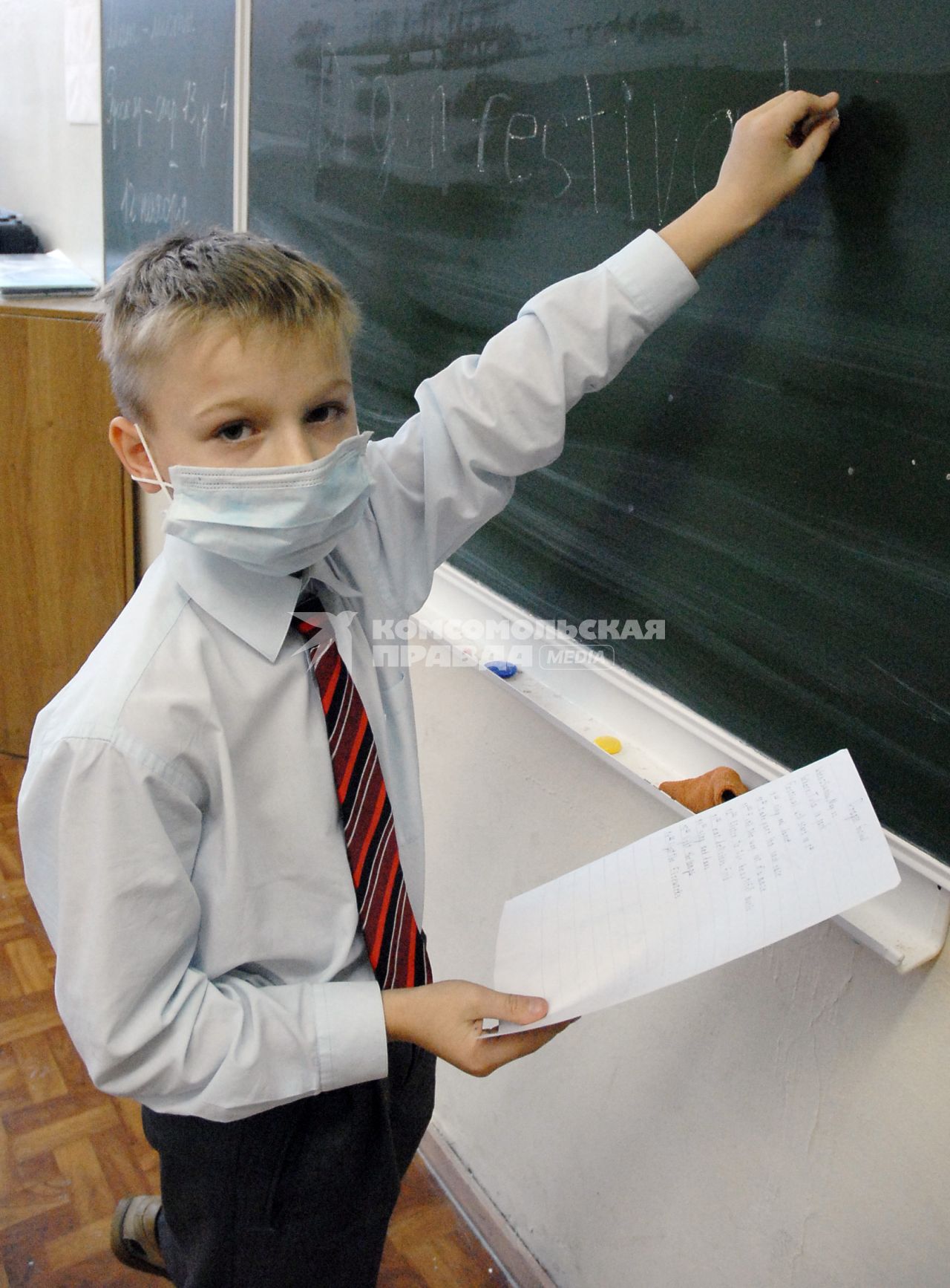 Школьник в медицинской повязке пишет мелом на доске.
