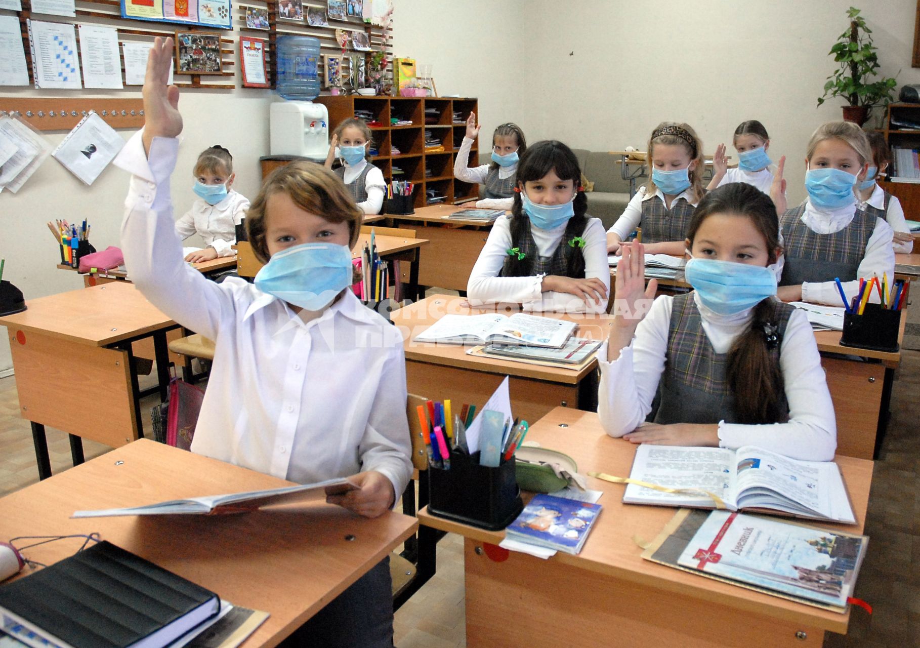 Школьники в медицинских повязках поднимают руки на уроке.