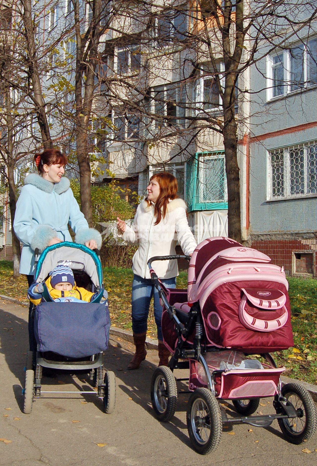 Две молодые мамы, гуляющие с колясками, общаются друг с другом на осенней улице на фоне жилого дома.