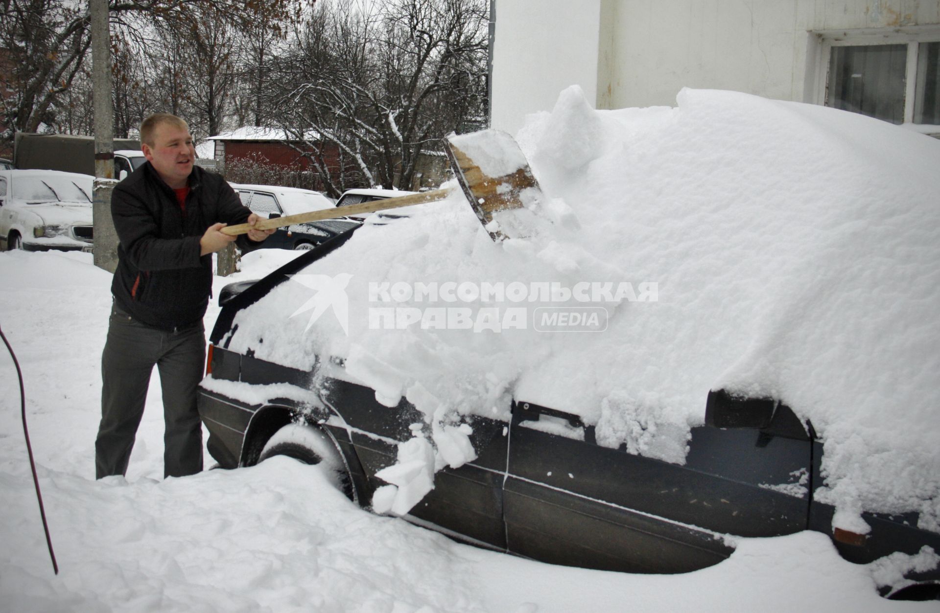 Мужчина отчищает автомобиль от снега с помощью деревянной лопаты.