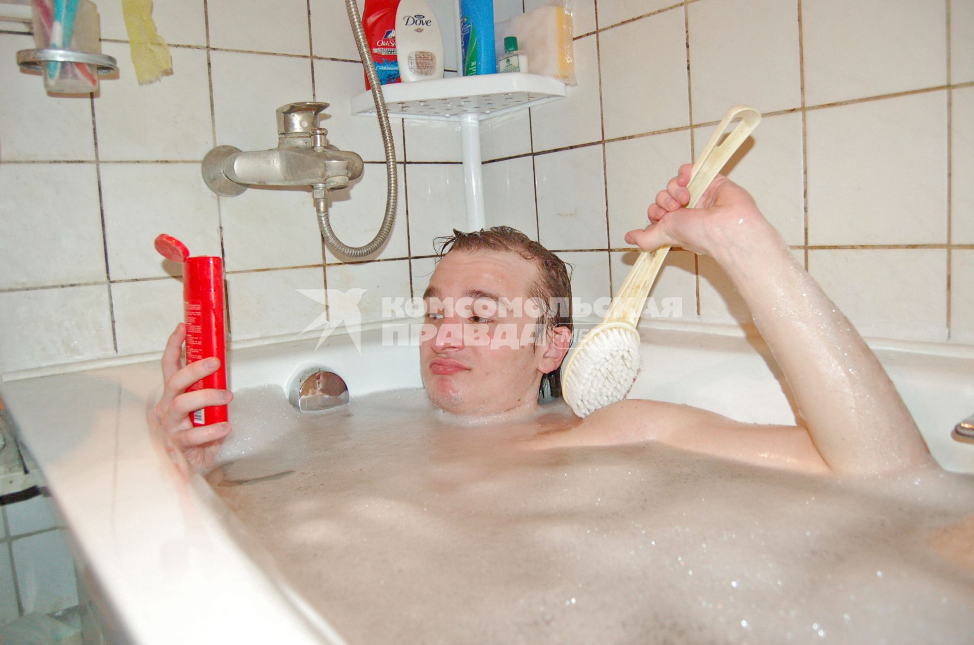 Парень, лежащий в ванне с пеной, держит флакон с моющим средством и щетку.