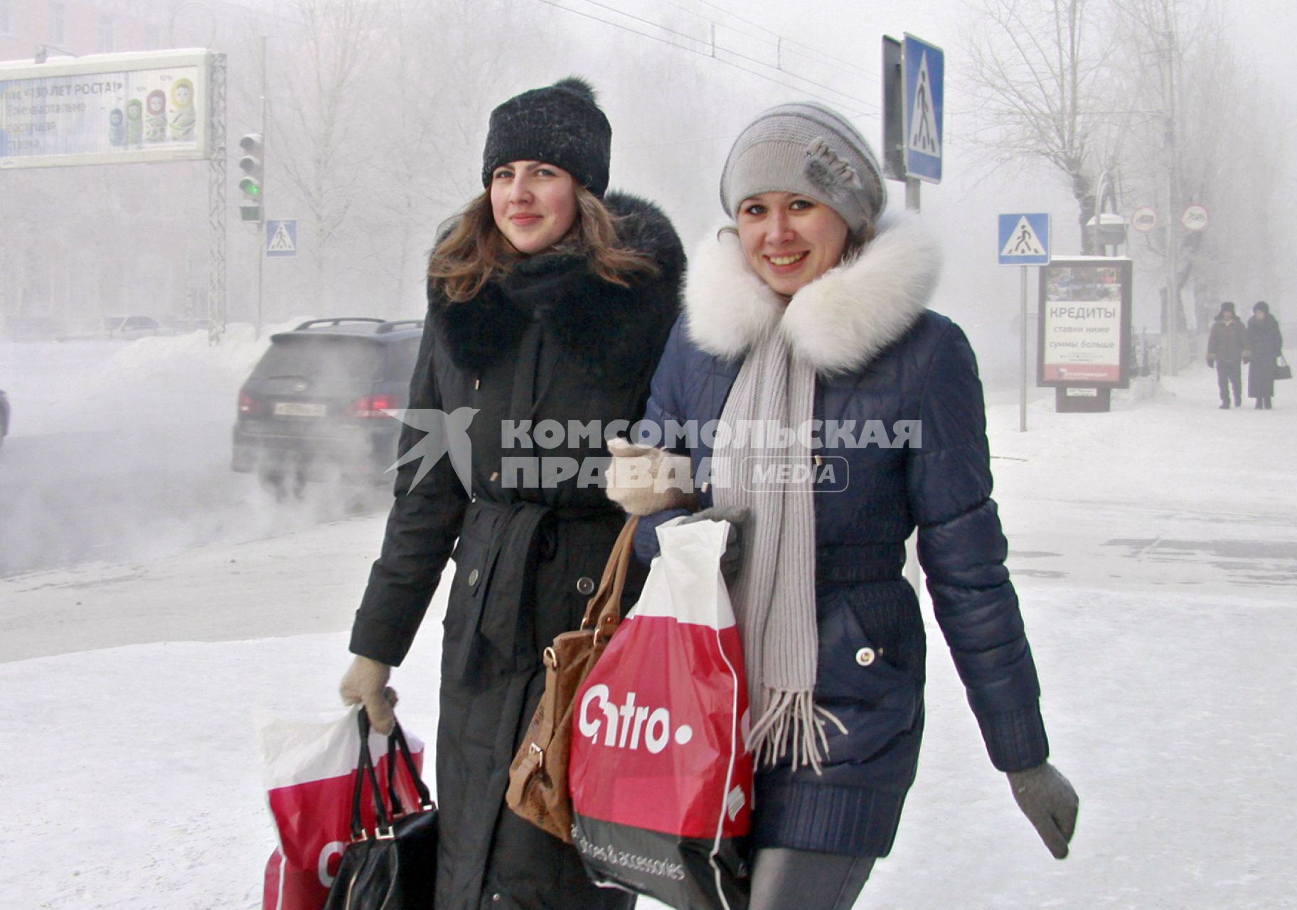 Две девушки улыбаясь идут под руку по заснеженной улице Барнаула.