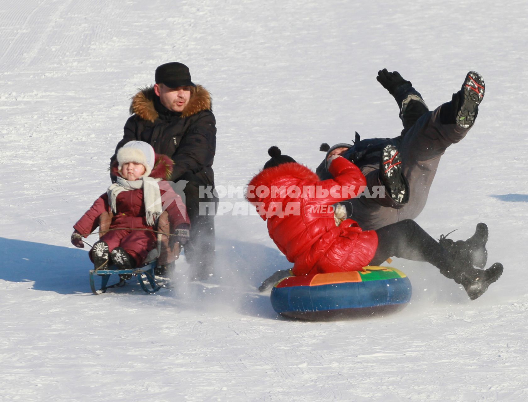 Девочка на тюбинге катаясь со снежной горки сбивает стоявшего мужчину. Рядом едет ребенок на санках, которого держит мужчина.