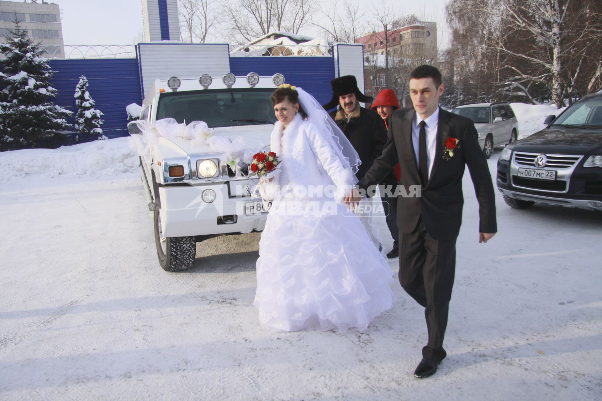 Молодожены зимой на улице идут мимо свадебного автомобиля.