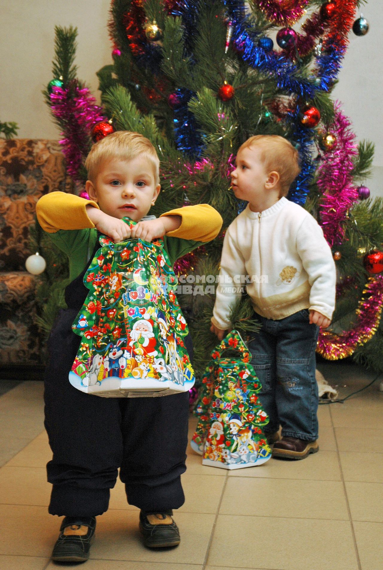 Ребенок держит в руках новогодний подарок с конфетами, второй ребенок со своим подарком стоит сзади.