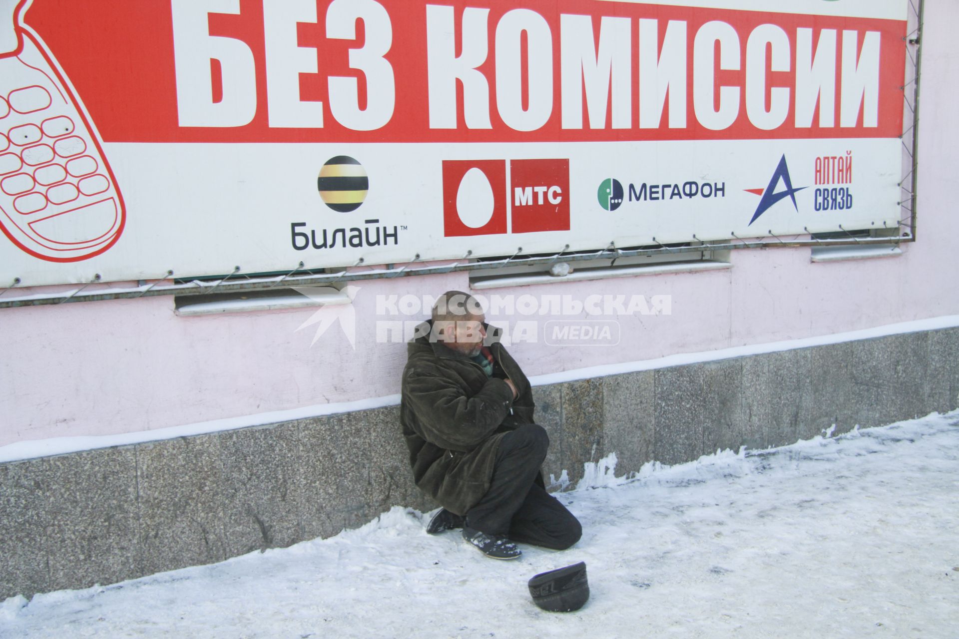 Бомж сидит зимой на снегу около рекламного плаката с надписью: `Без комиссии`.