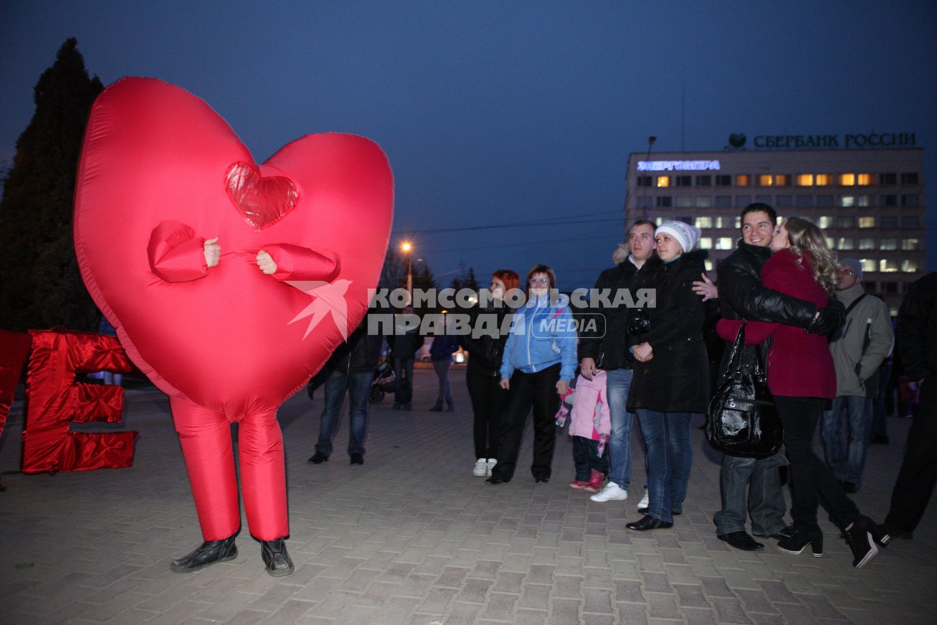 Человек в костюме сердечка на улице среди людей.