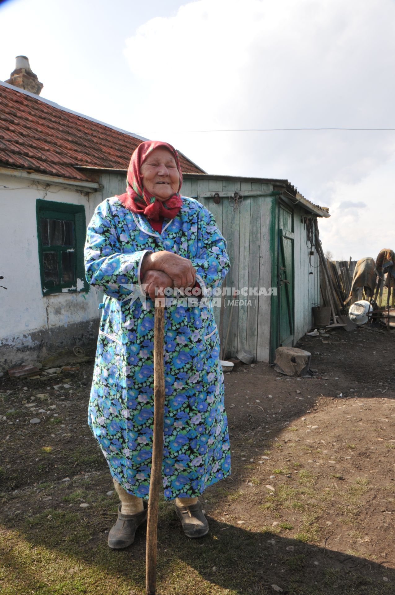 Пенсионер стоит рядом со своим домом в деревне.