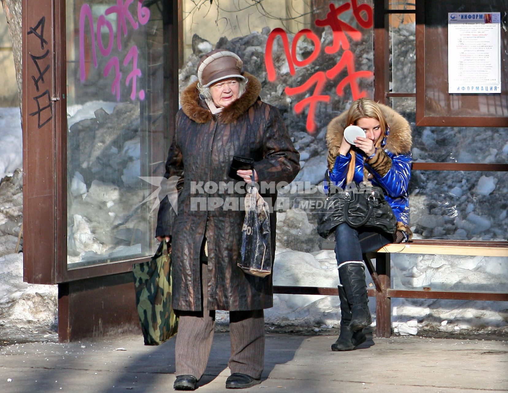 Девушка подкрашивает глаза на автобусной остановке, рядом стоит пожилая женщина.