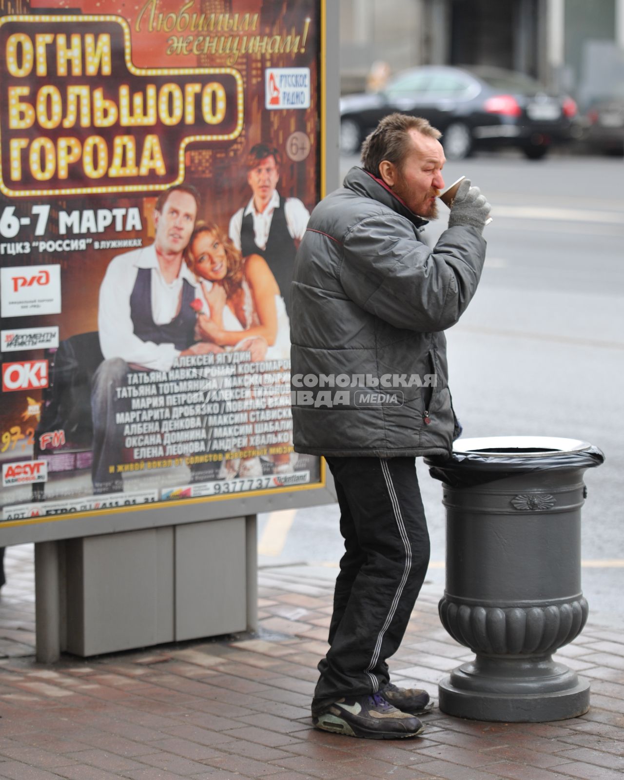 Бомж пьет кофе на улице рядом с урной.