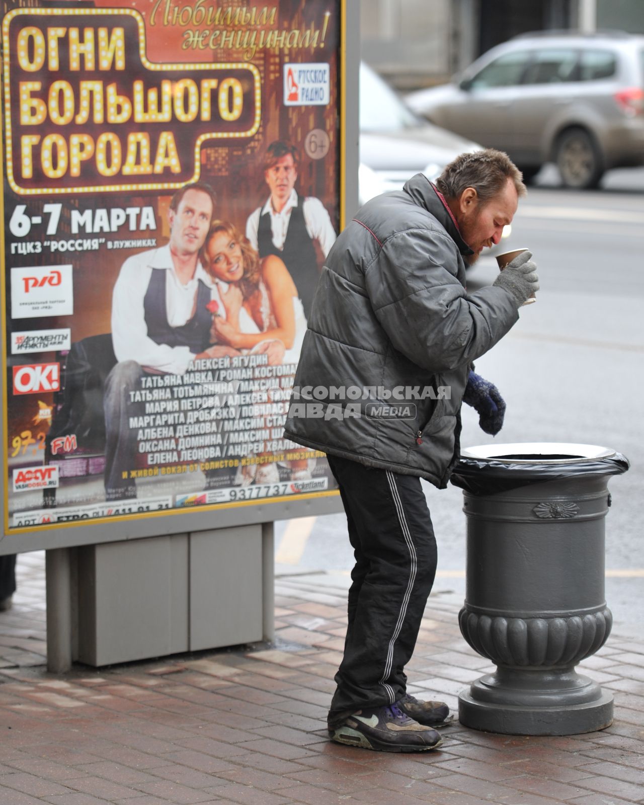 Бомж пьет кофе на улице рядом с урной.