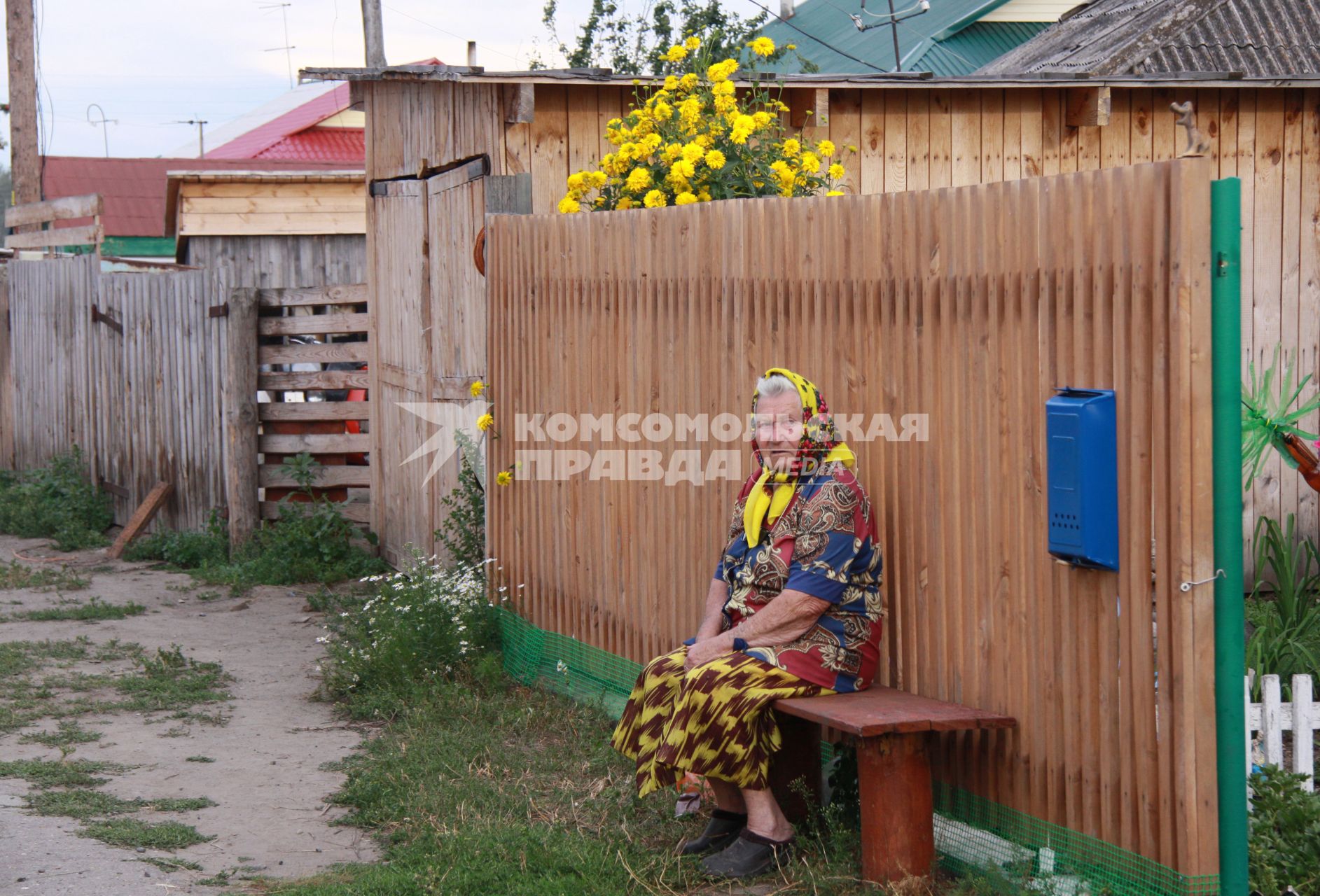 Бабушка сидит на лавочке перед сельским домом.