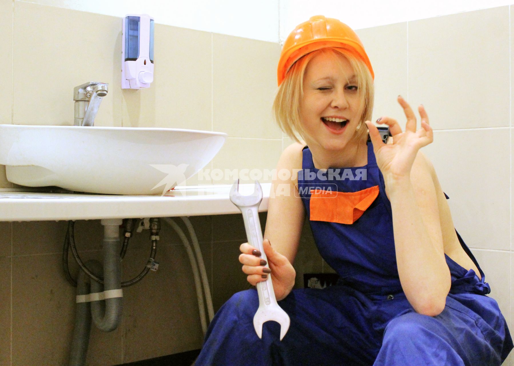 Девушка в спецодежде и каске занимается ремонтом водопроводного крана в ванной комнате.