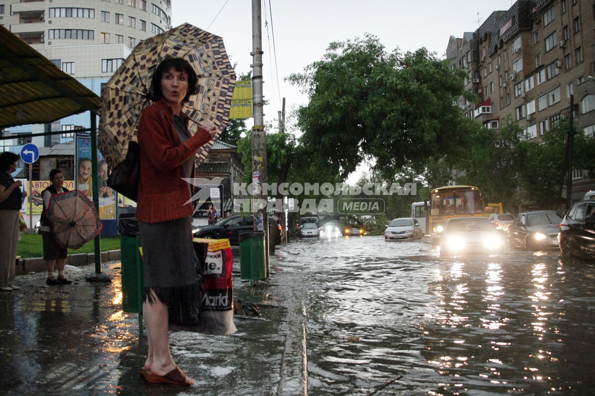 Женщина с зонтом стоит на остановке около затопленной улицы после сильного ливня.