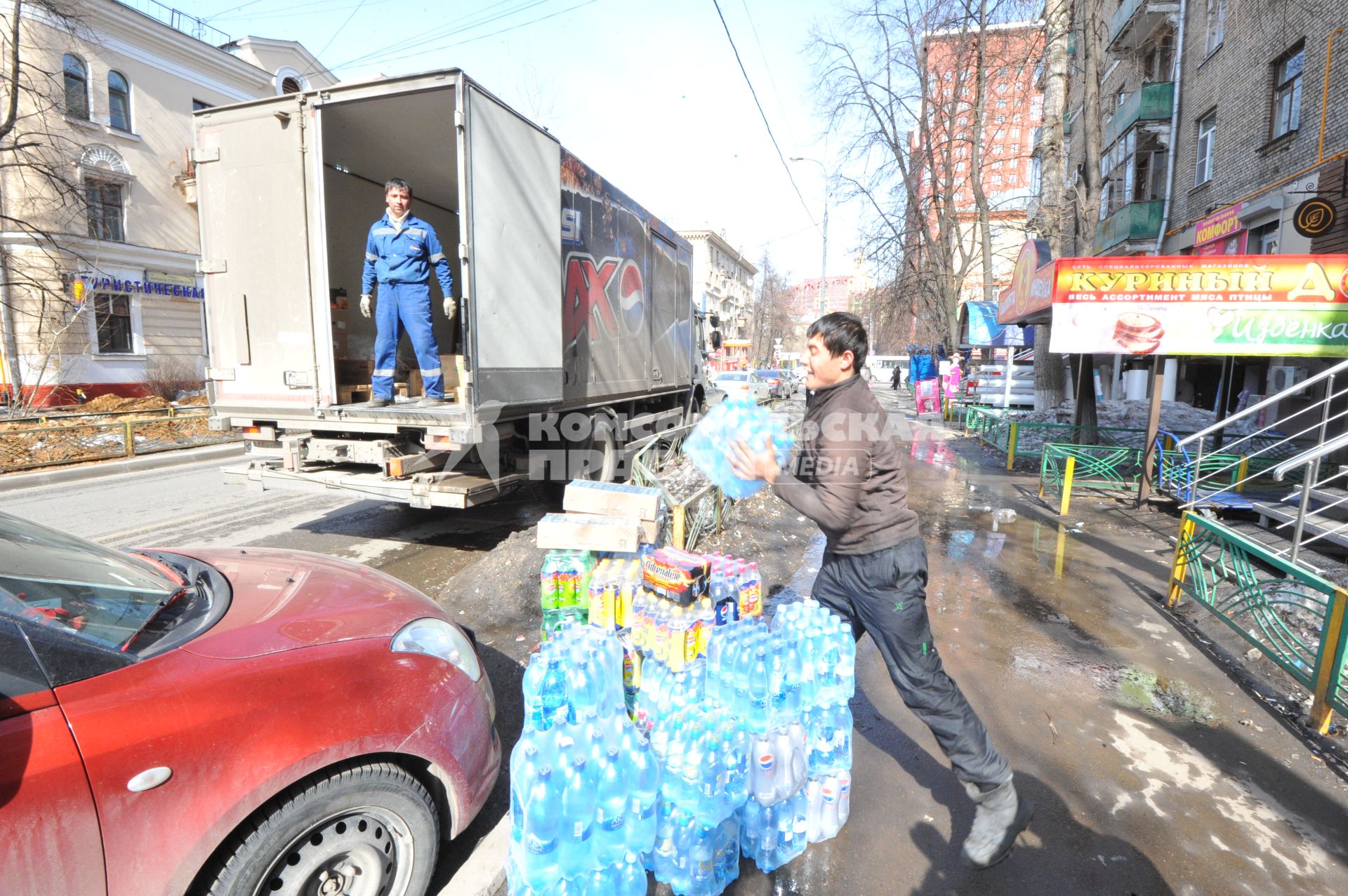 Рабочие выгружают из машины упаковки с водой и напитками. Магазин `Куриный дом` (справа).