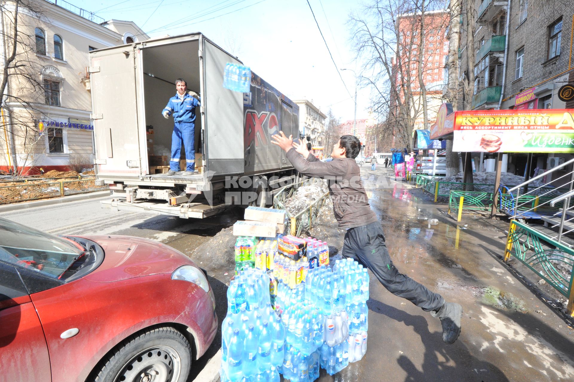 Рабочие выгружают из машины упаковки с водой и напитками. Магазин `Куриный дом` (справа).