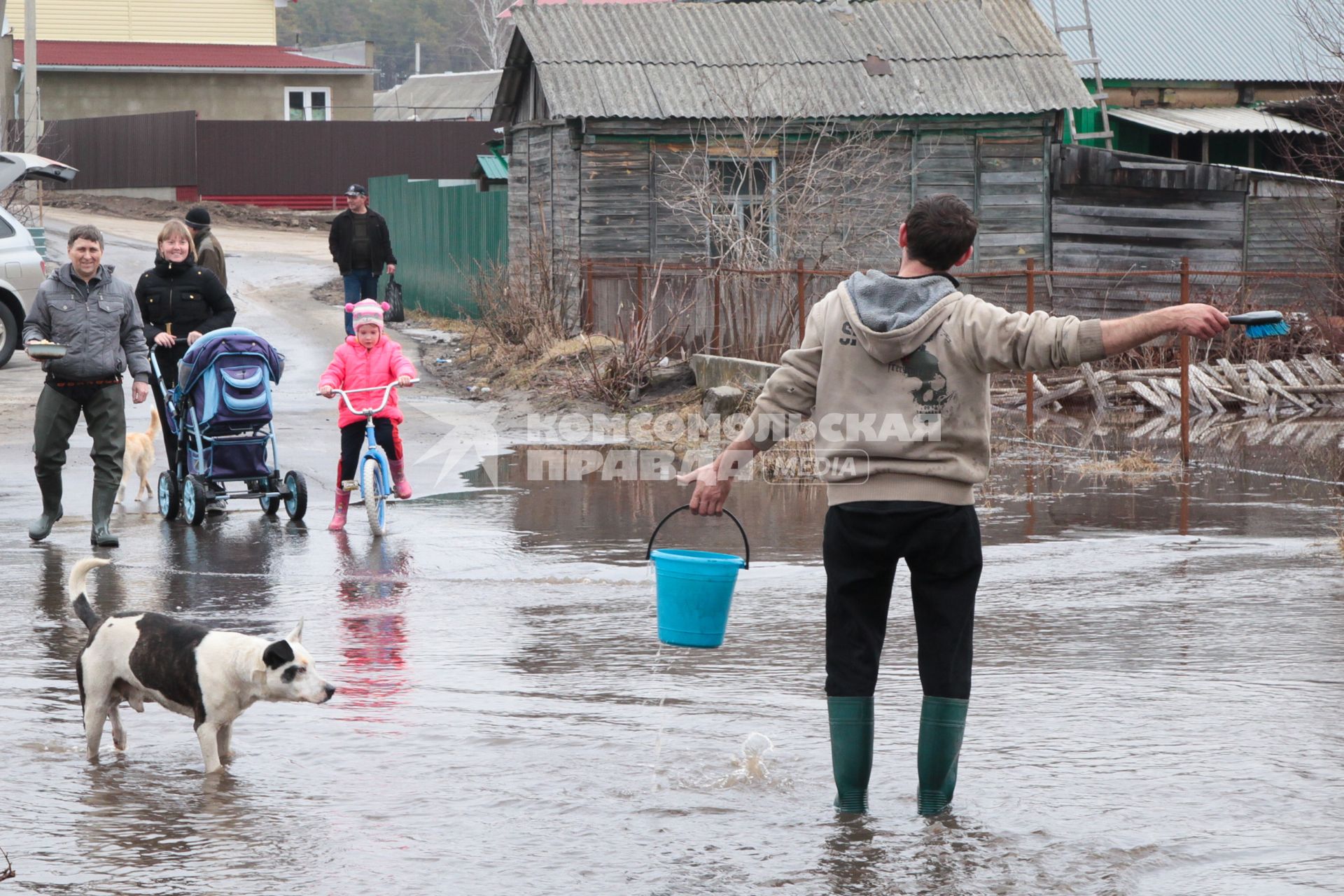 Мужчина с ведром воды идет по затопленной улице, на него смотрят жители села.