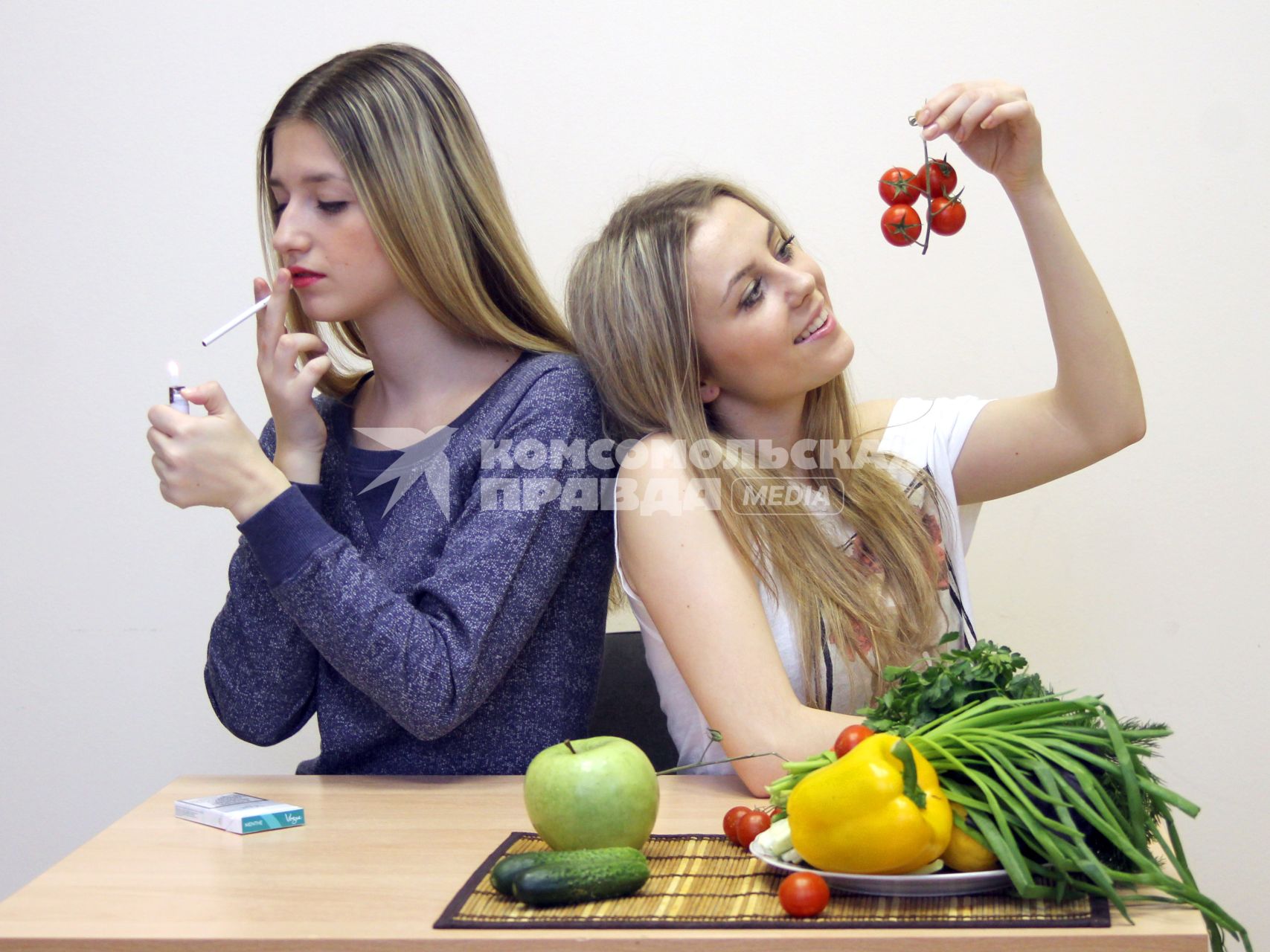 Девушки стоят за столом с овощами и фруктами. Одна держит в руках томаты, другая - сигарету.