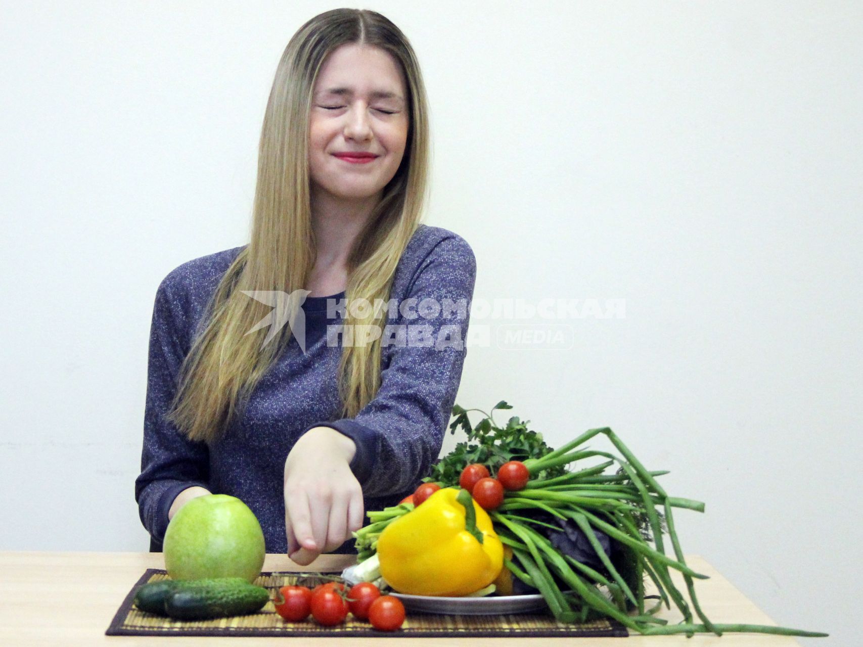 Девушка сидит за столом с овощами и фруктами зажмурившись.