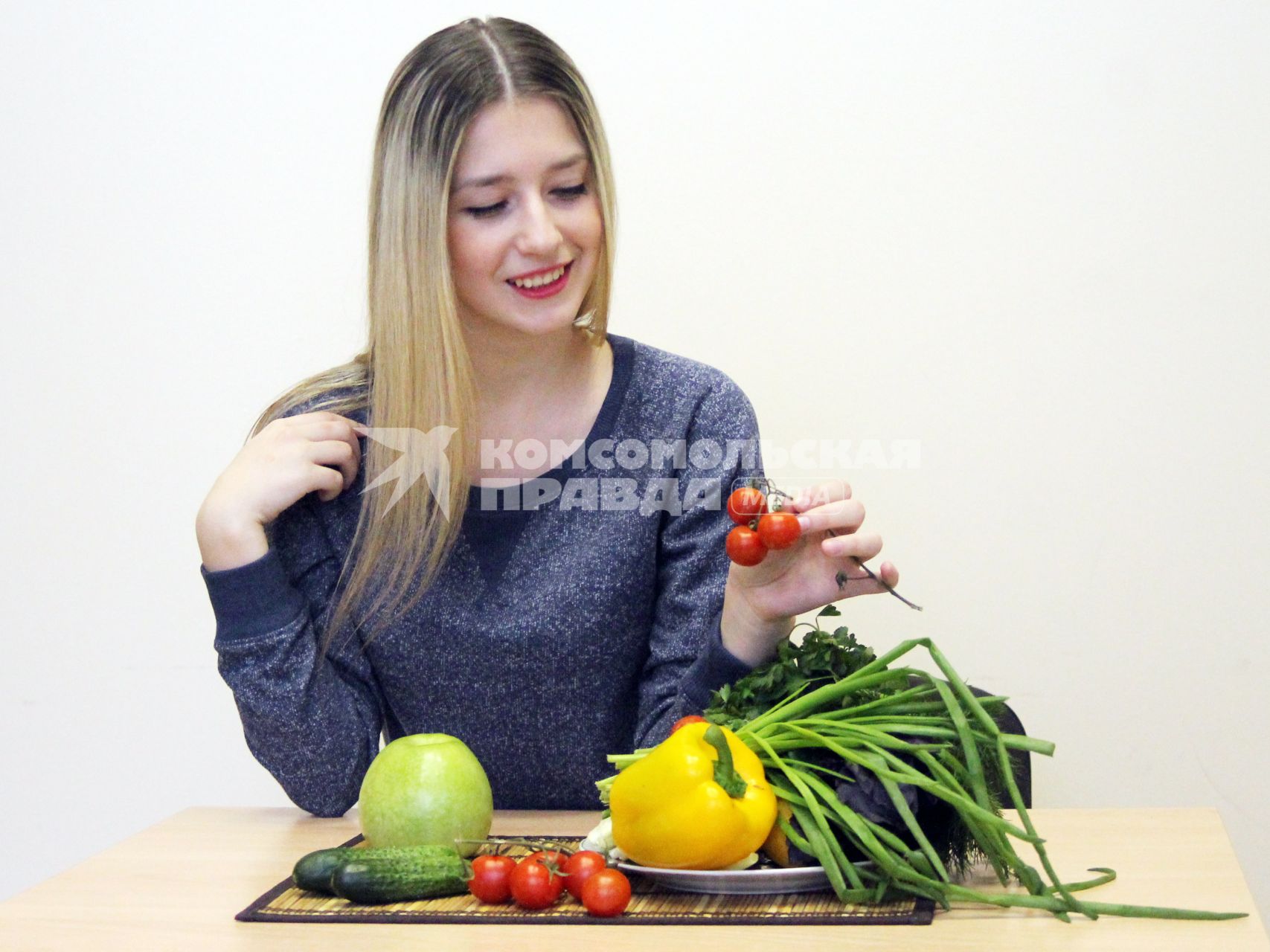 Девушка сидит за столом с овощами и фруктами и держит в руках томат.
