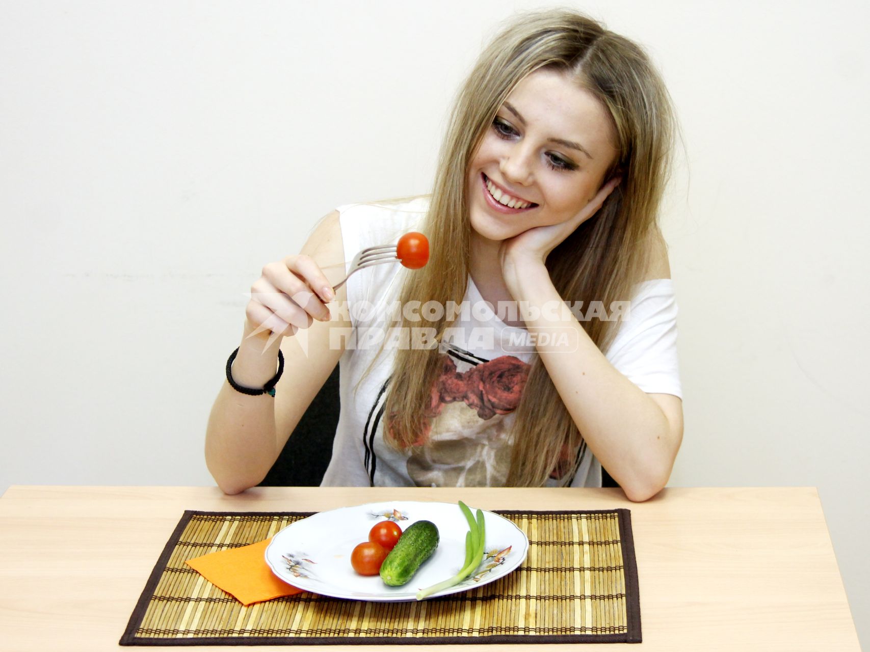 Девушка сидит за столом перед тарелкой с овощами и кушает томат.