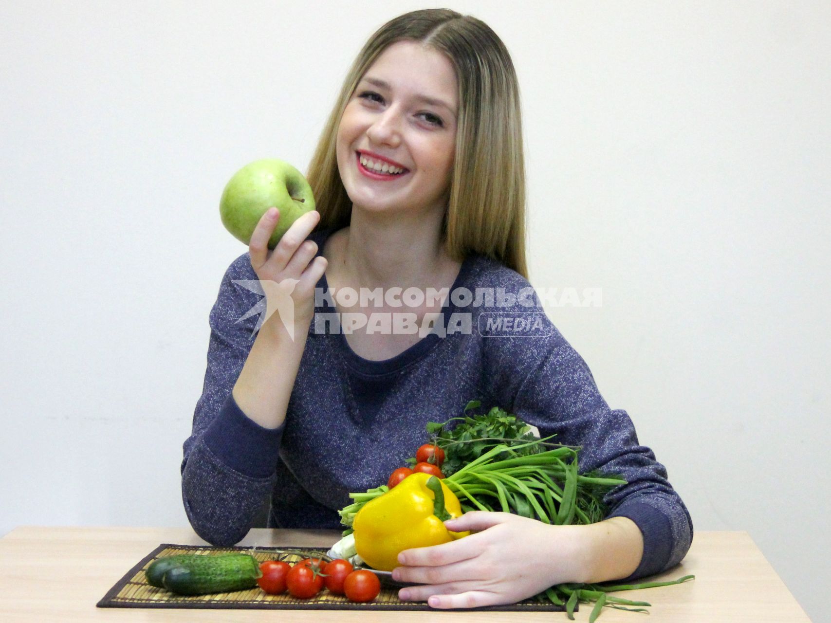 Девушка сидит за столом с овощами и фруктами и держит в руках яблоко.