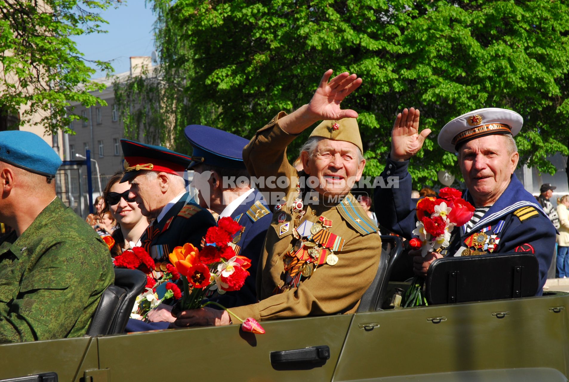 Ветераны в парадной форме и с цветами приветственно машут из открытой военной машины.