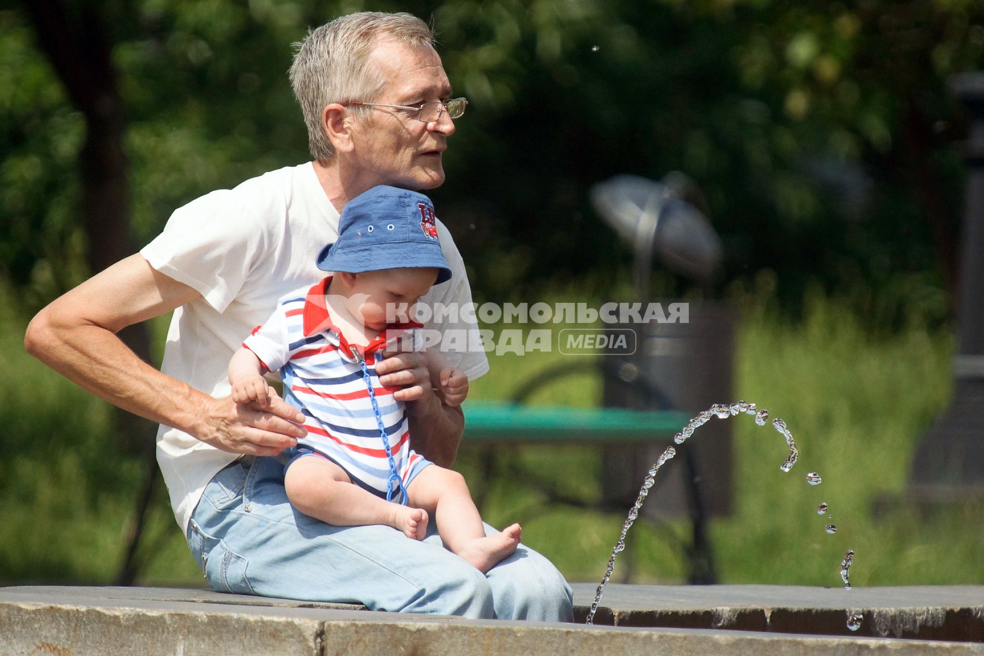 Ребенок, сидя на коленках у деда, наблюдает за струей воды в фонтане.