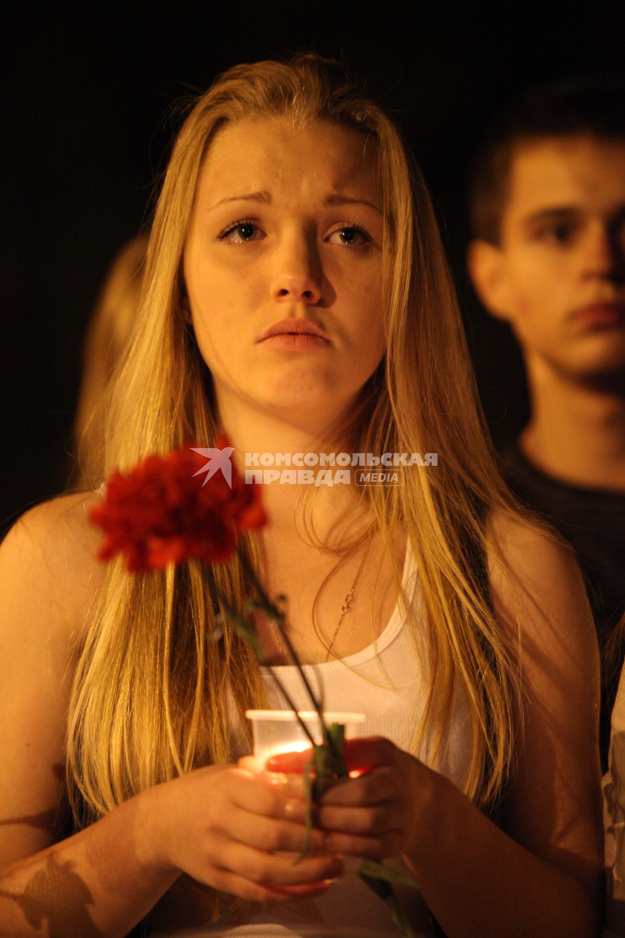 Молодая девушка держат в руках стаканчик со свечой и гвоздики, в глазах у нее слезы