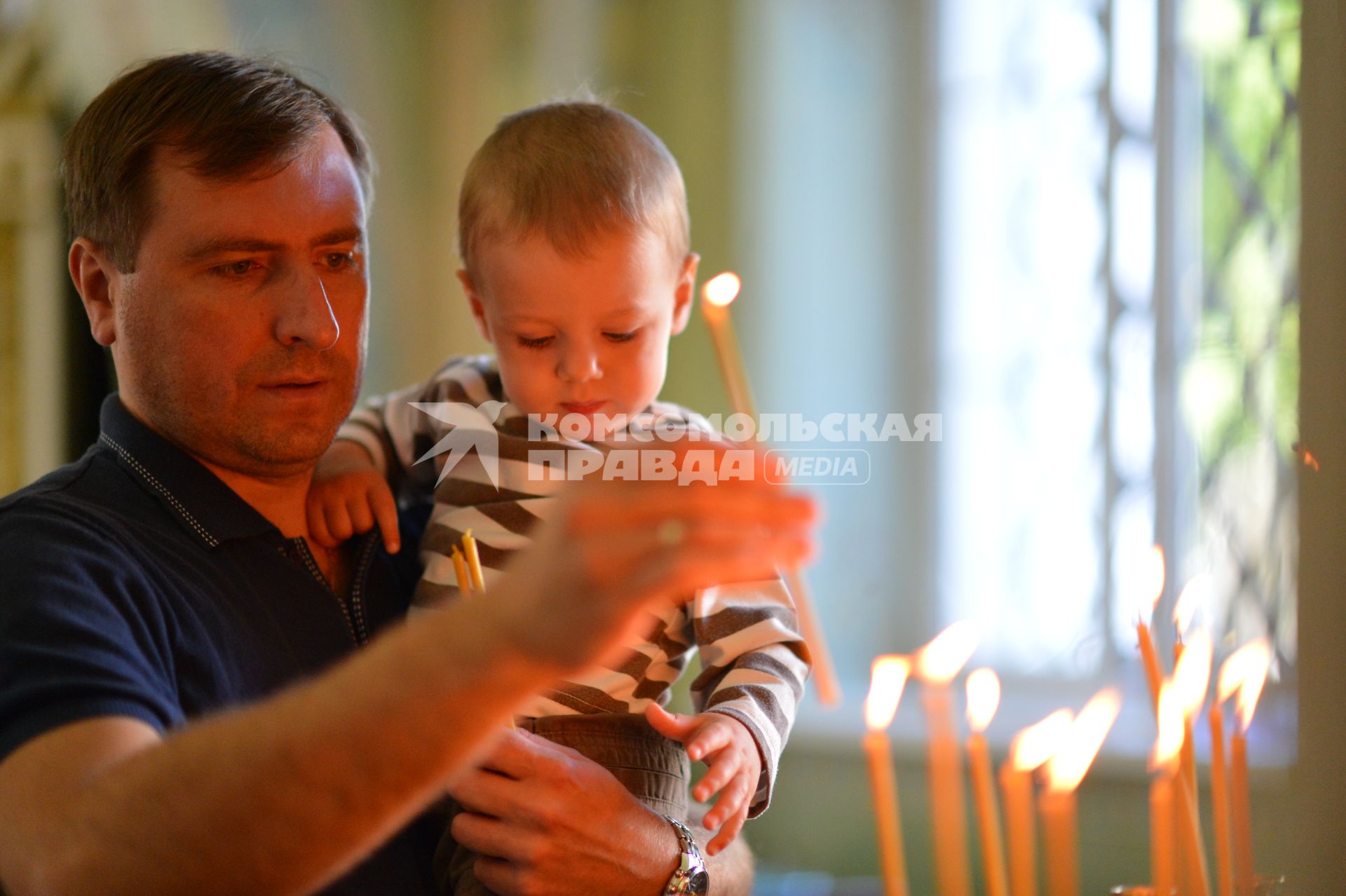 Мужчина с ребенком на руках ставить свечу в подсвечник.
