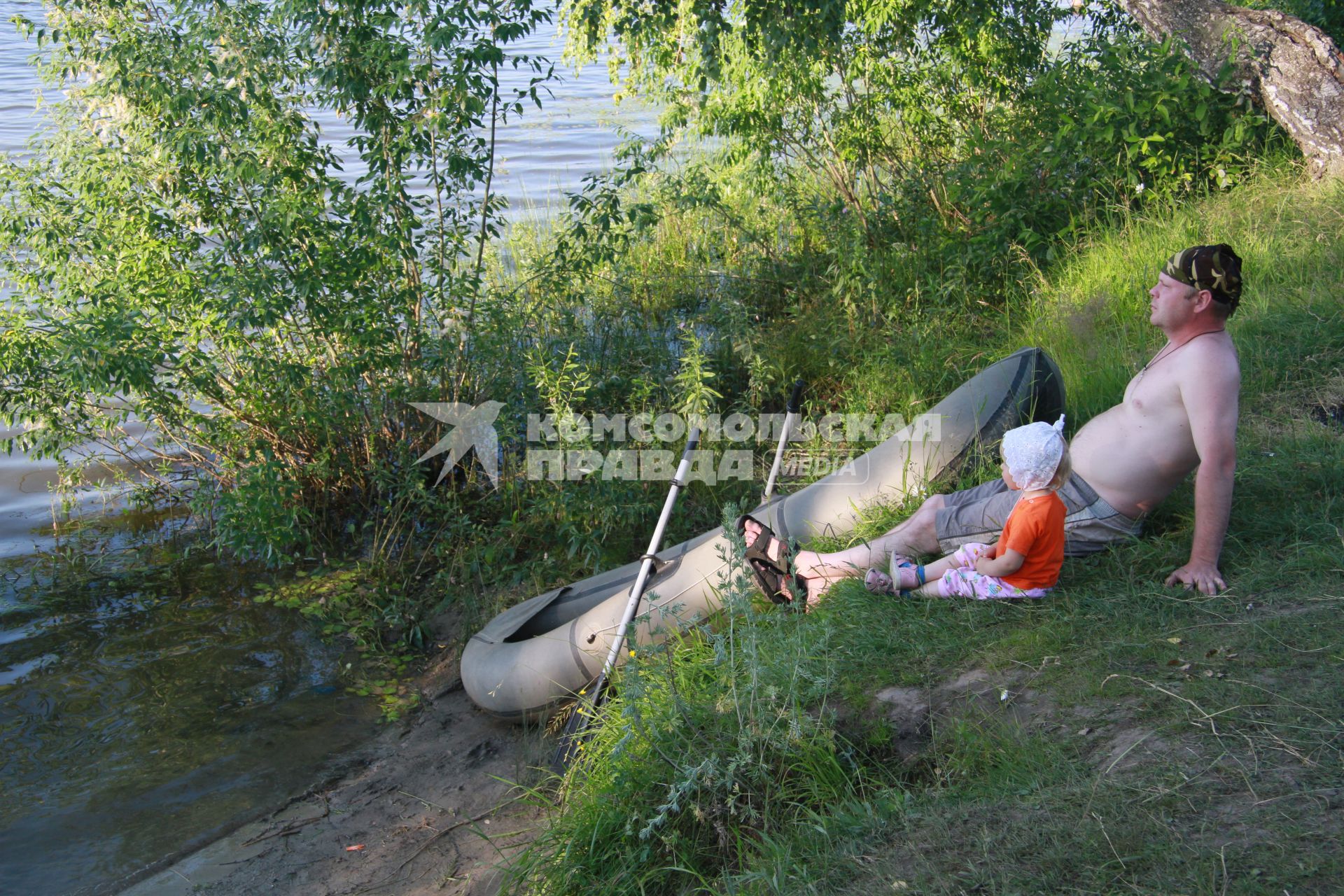 Мужчина с маленькой девочкой сидят на берегу водоема с надувной лодкой.