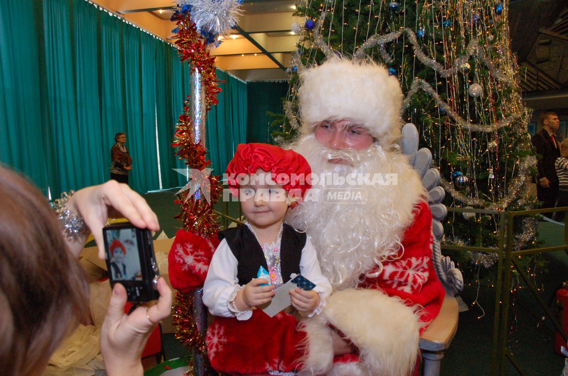 Дети и родители в карнавальных костюмах на Новогодней елке. Девочка в костюме красной шапочки сидит на коленях у Деда Мороза.