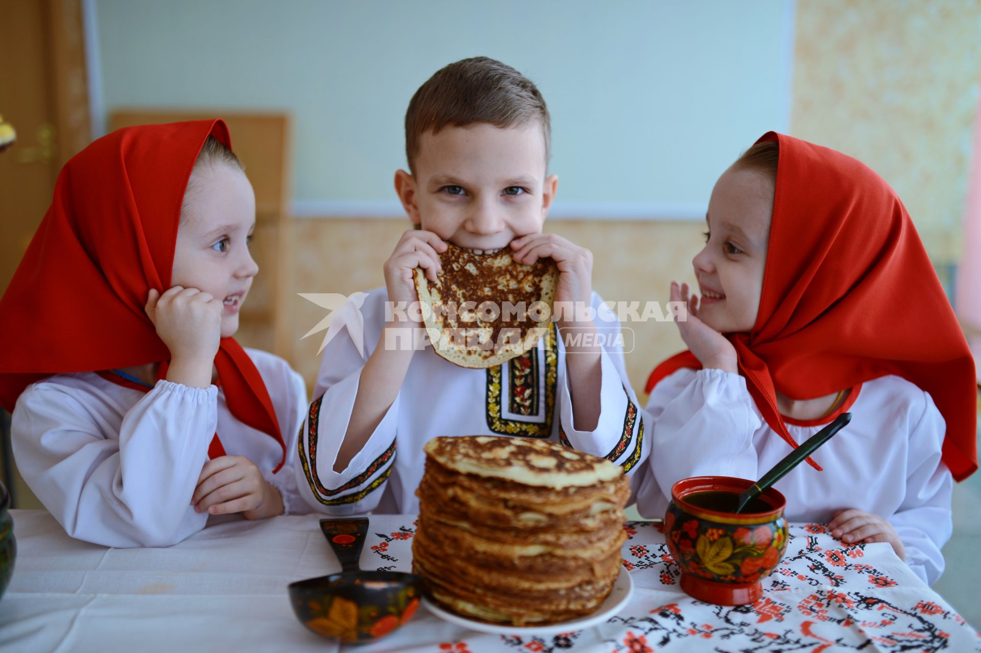 Масленица. На снимке: девочки-близняшки и мальчик в русских народных костюмах сидят за столом с блинами.