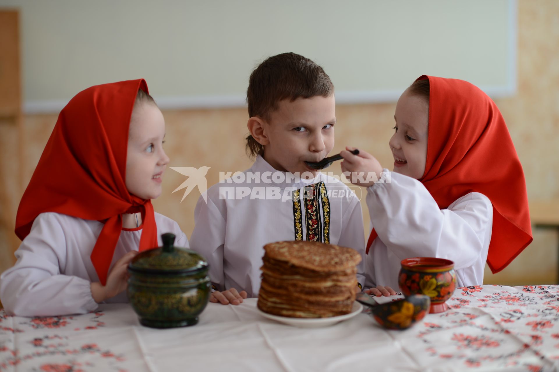 Масленица. На снимке: девочки-близняшки и мальчик в русских народных костюмах сидят за столом с блинами.
