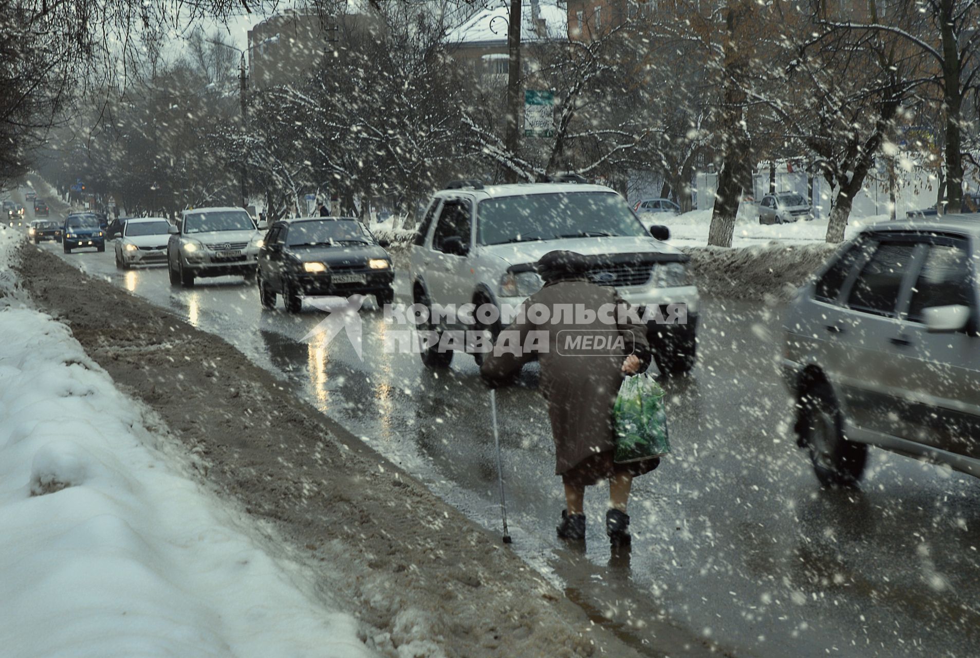 Пенсионерка с палочкой бредет под снегопадом по обочине навстречу едущим автомобилям.