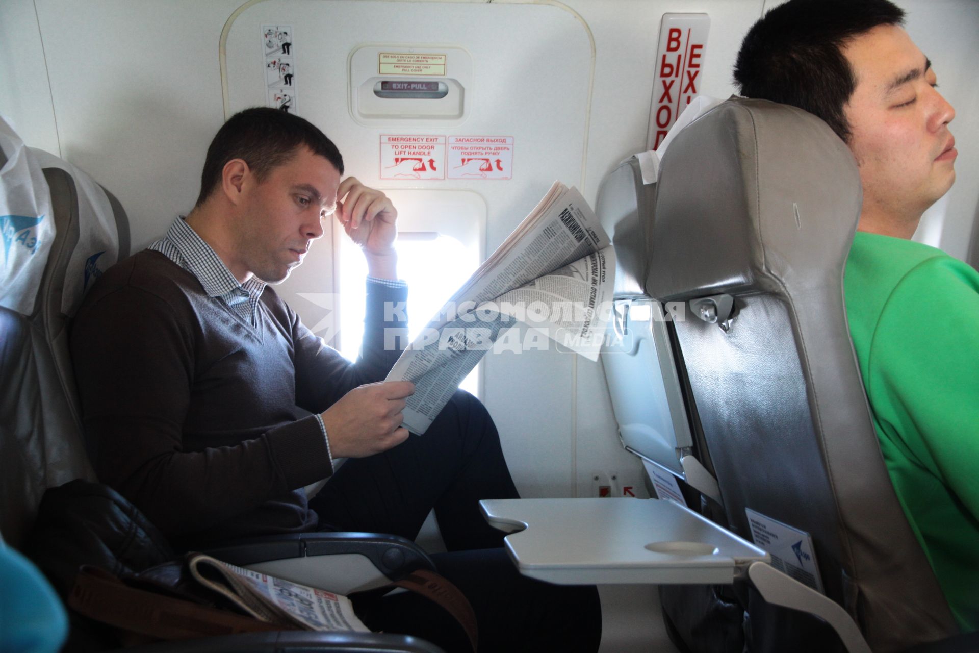 Мужчина читает газету в салоне самолета сидя у запасного аварийного выхода.