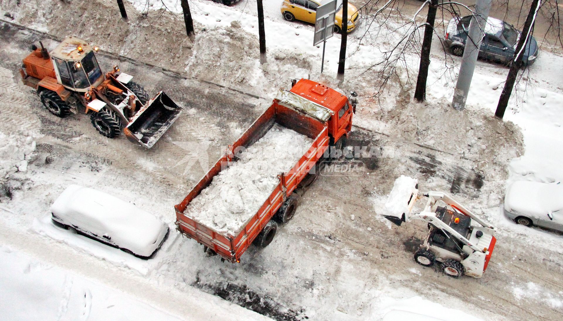 Трактор и минипогрузчик загружают снег в грузовкик `Камаз`.