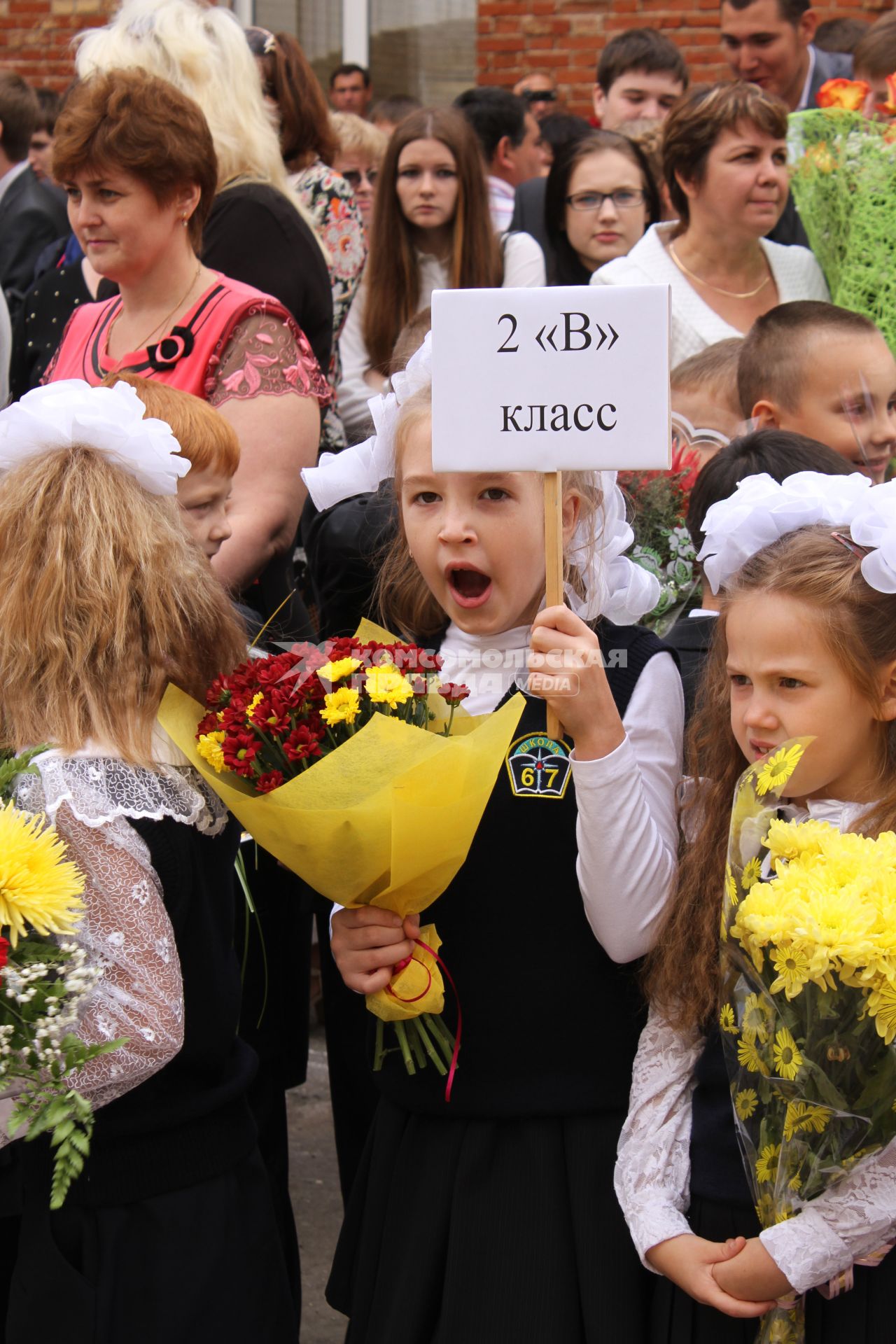 Зевающая девочка с табличкой `2 `Б` класс` на утренней школьной линейке.