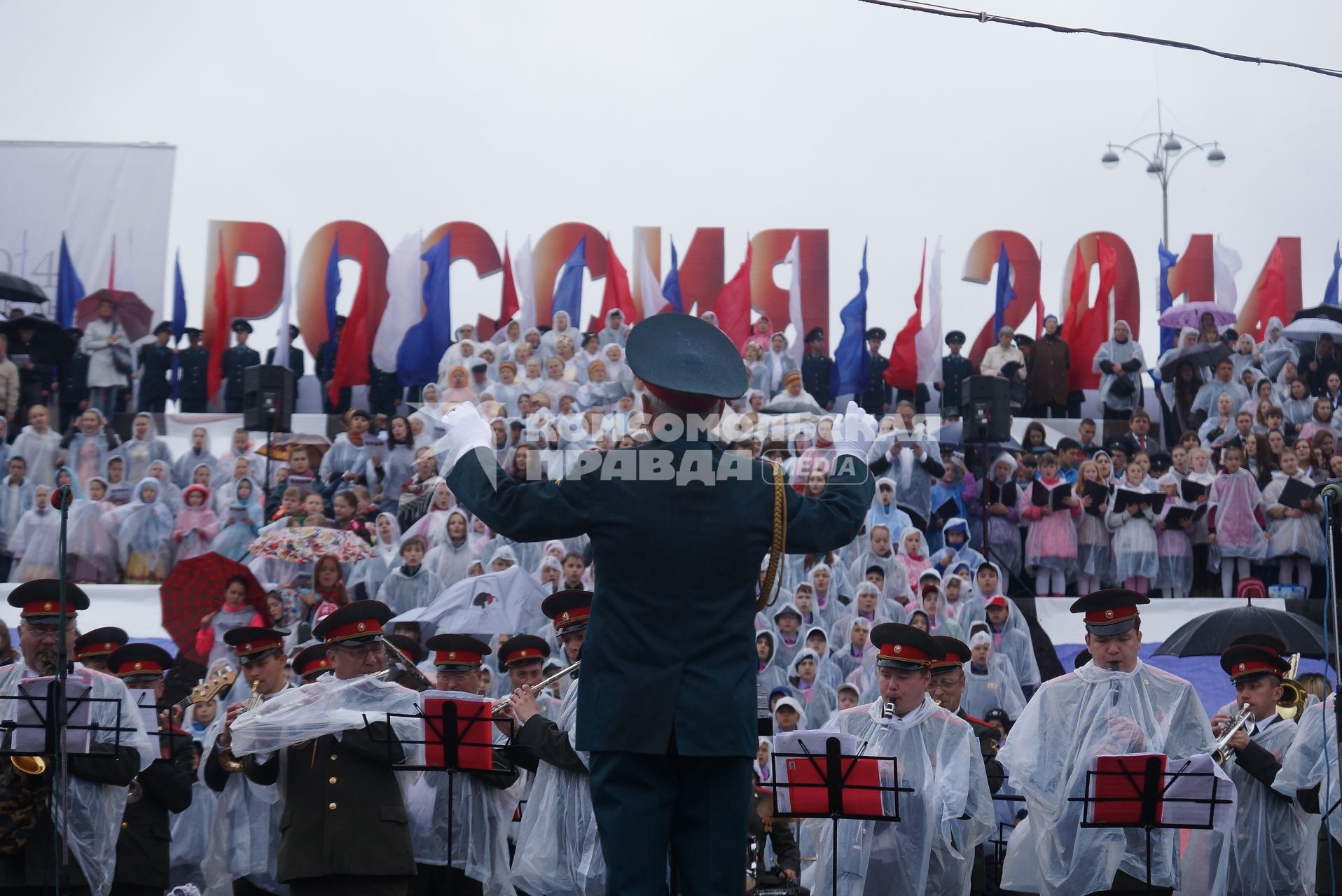 Дирижер под дождем руководит оркестром и сводным зором во время выступления на праздновании дня России