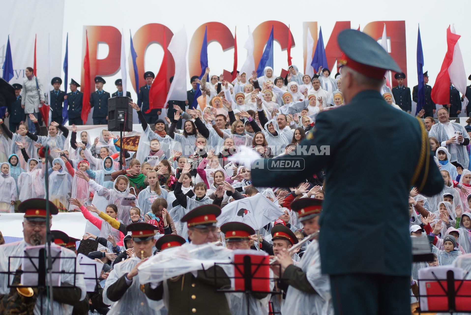 Дирижер под дождем руководит оркестром и сводным зором во время выступления на праздновании дня России