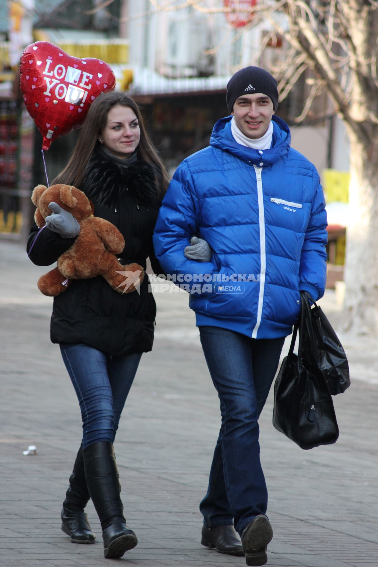 Мужчина и женщина идут подруку. У женщины в руках плюшевый медведь и надувной шар в виде сердца.