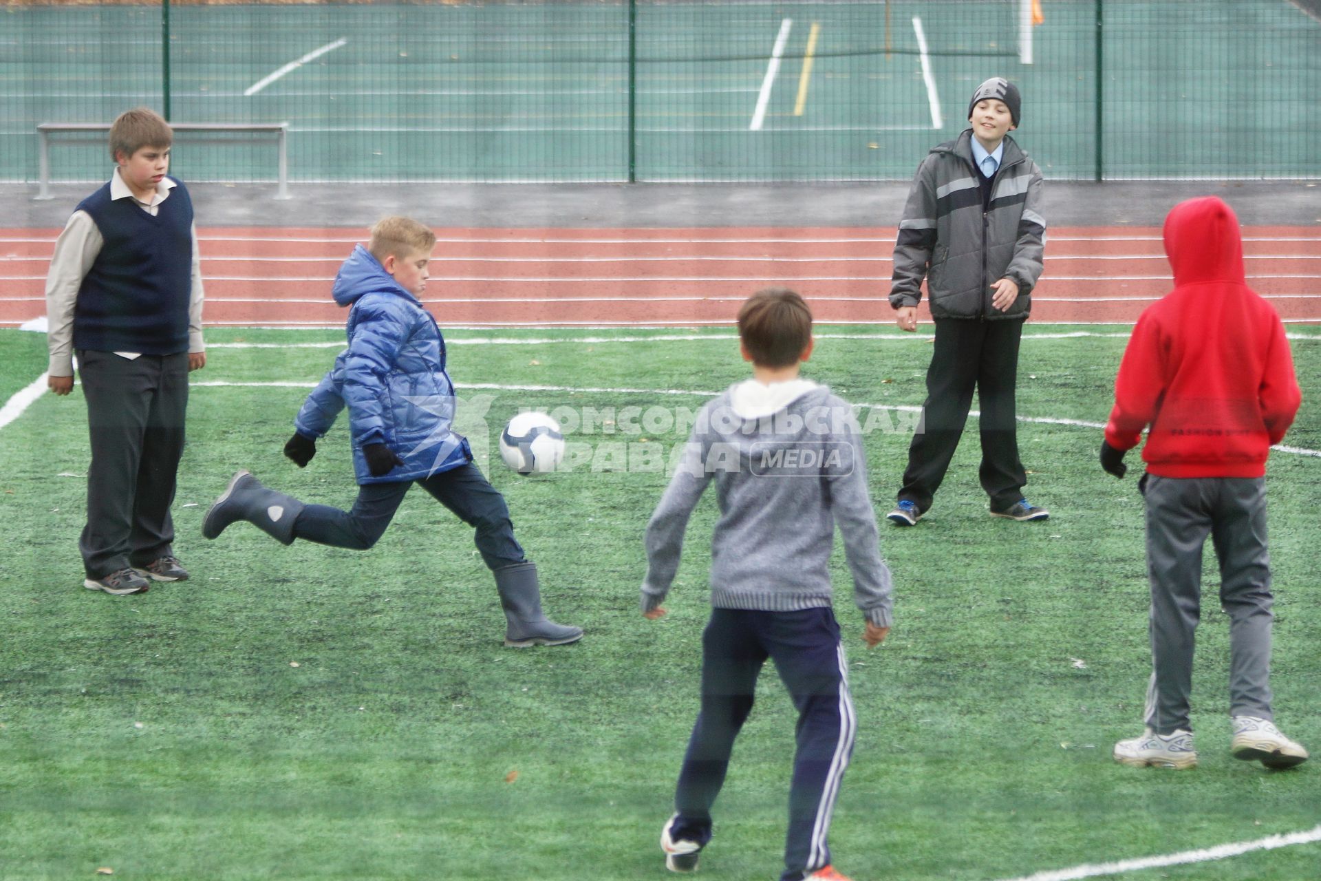 Мальчик в резиновых сапогах играет в футбол на школьном стадионе
