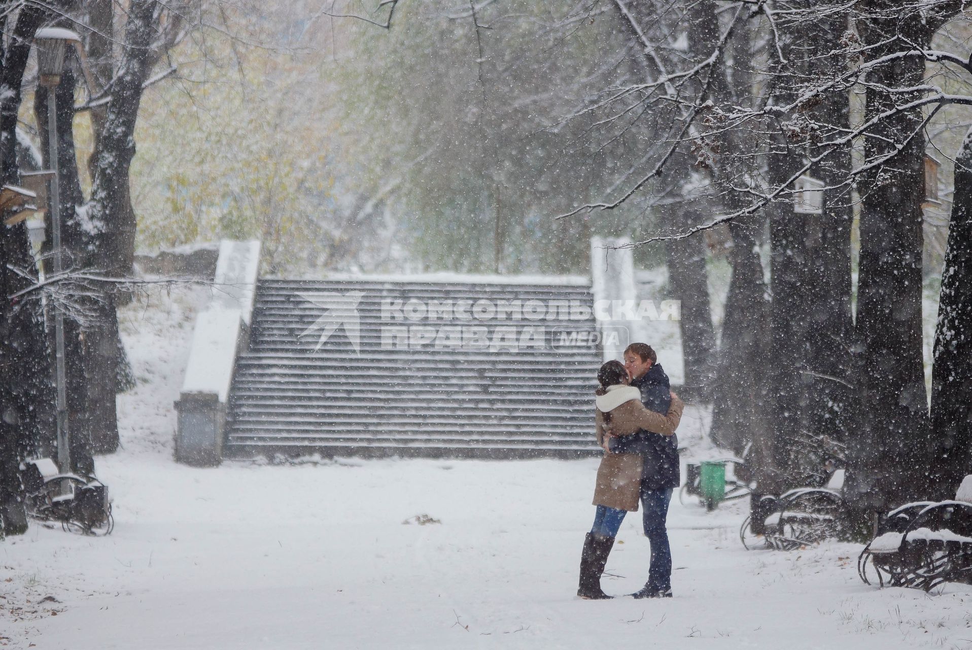 Влюбленные парень и девушка целуются в парке во время снегопада