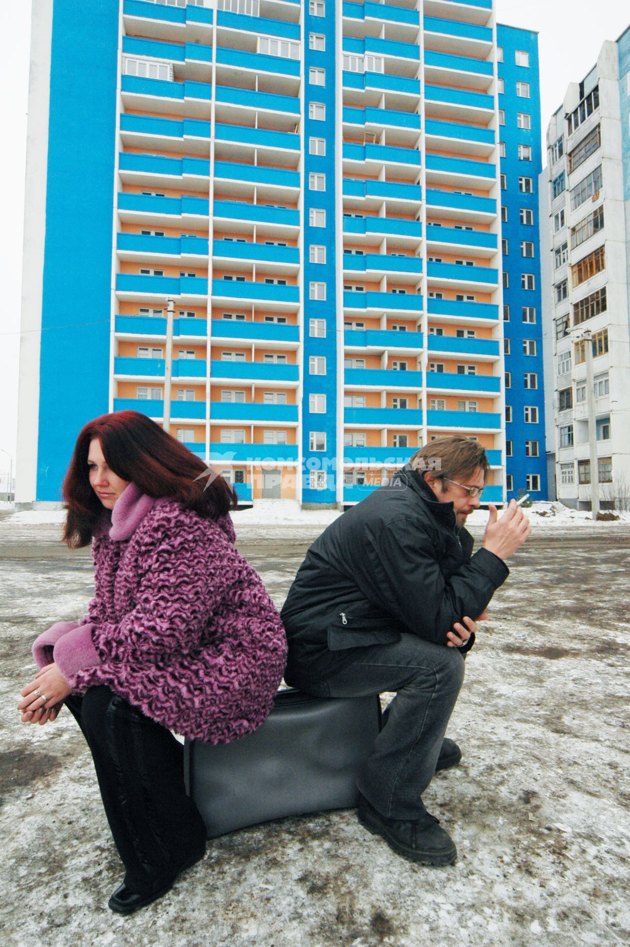 Женщина и мужчина сидят на чемодане на фоне нового многоквартирного дома.