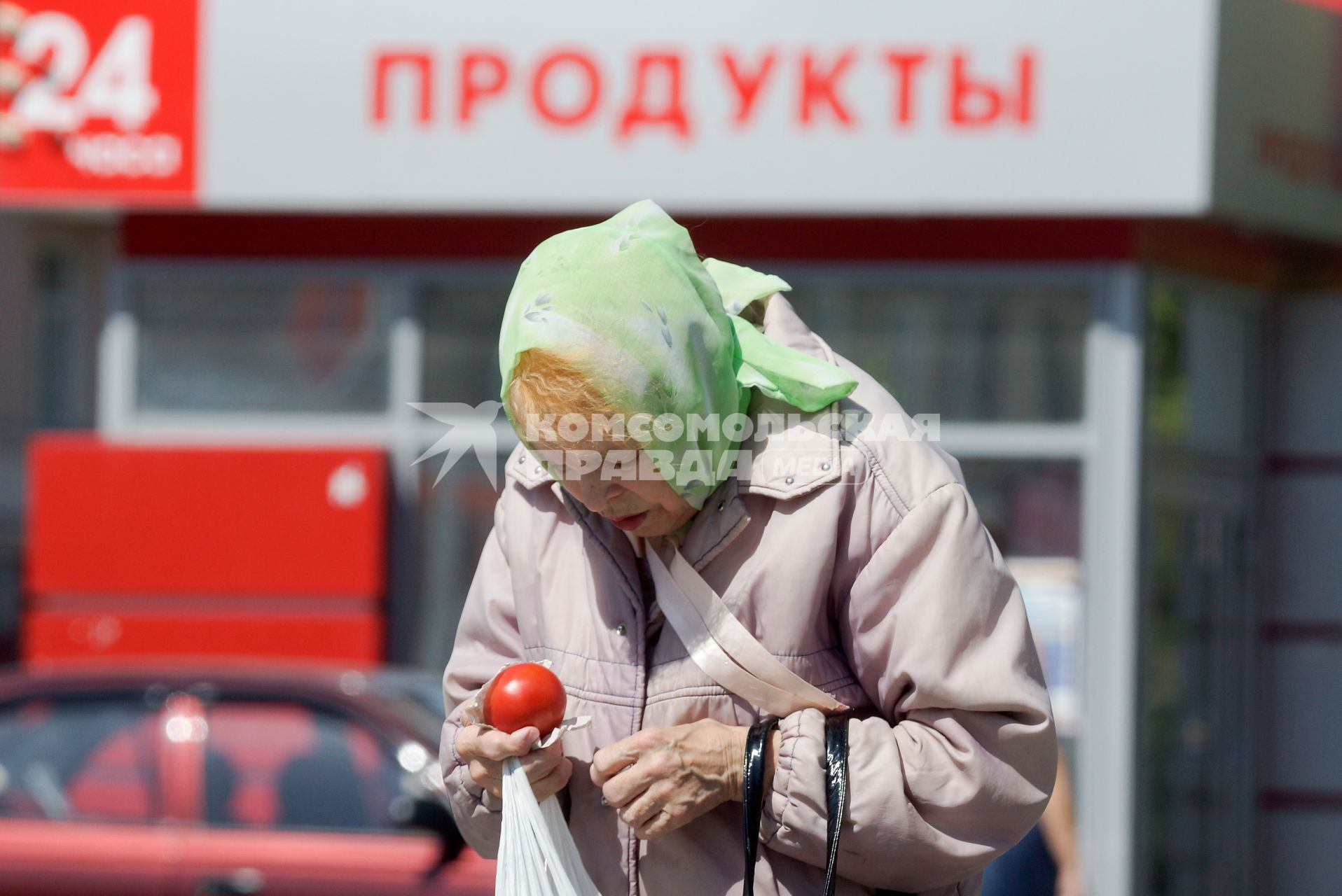 Бабушка держит в руках пакет с продуктами и помидор, у продуктового ларька