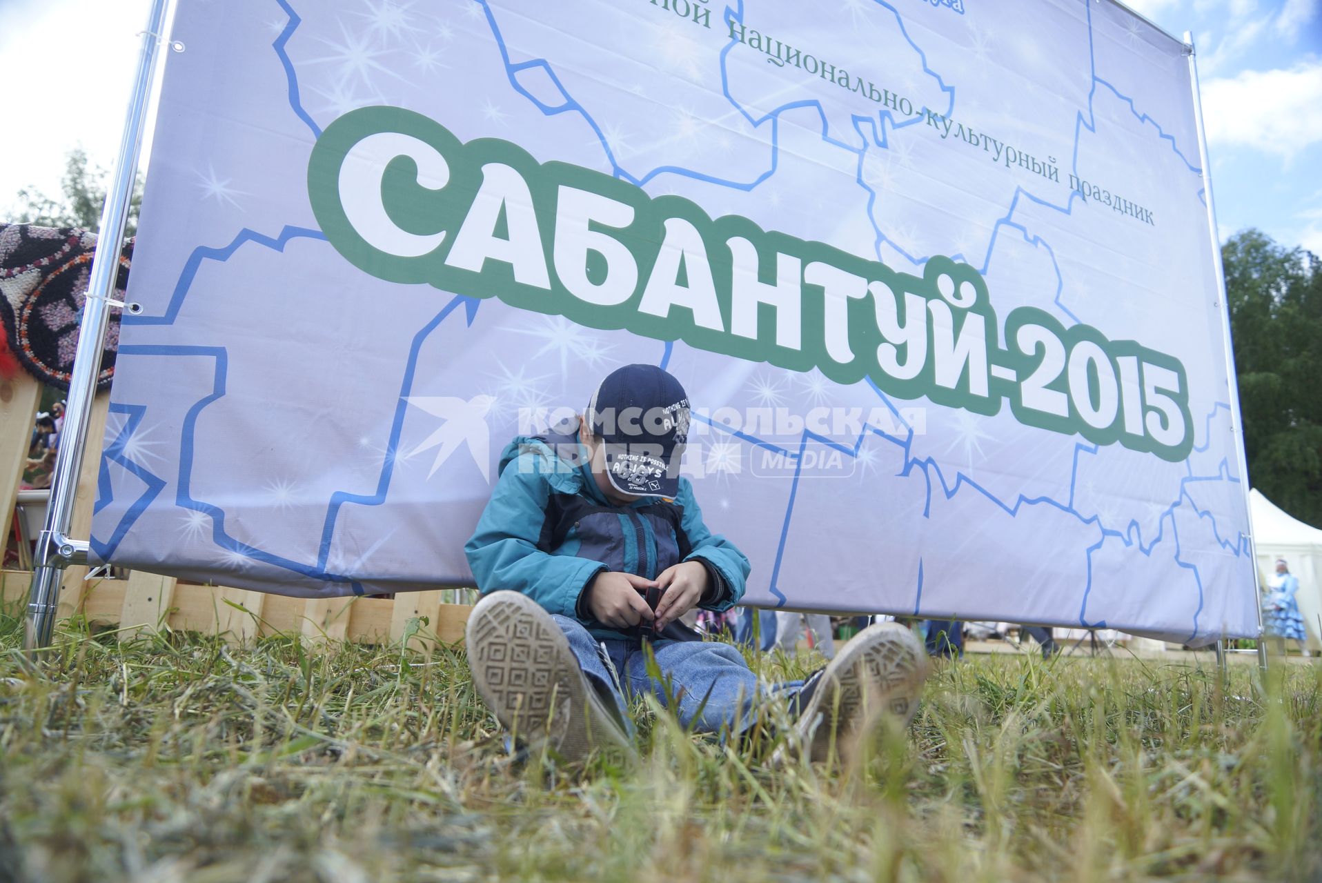 Ребенок сидит у плаката с надписью \"Сабантуй-2015\", во время празднования Сабантуя. село Кадниово. Свердловская область